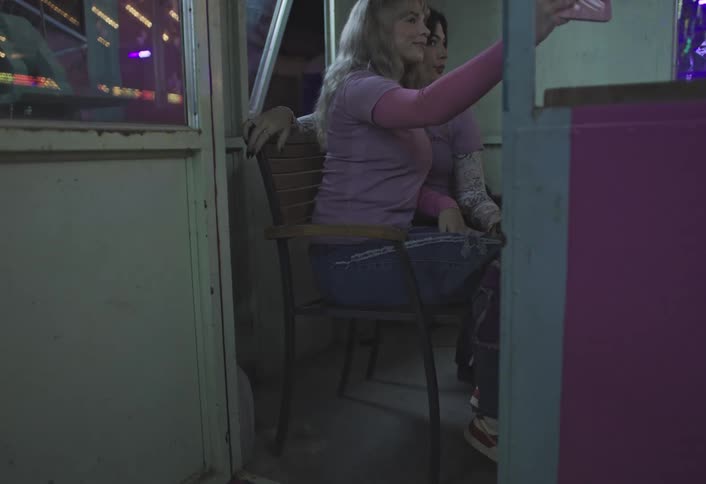 Two women sit in a booth taking selfies with a smartphone at an amusement park at night