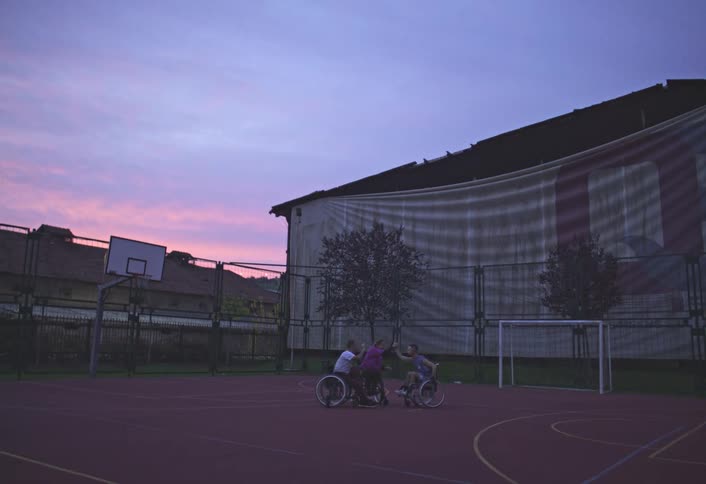 Three people in wheelchairs play basketball on an outdoor court during sunset
