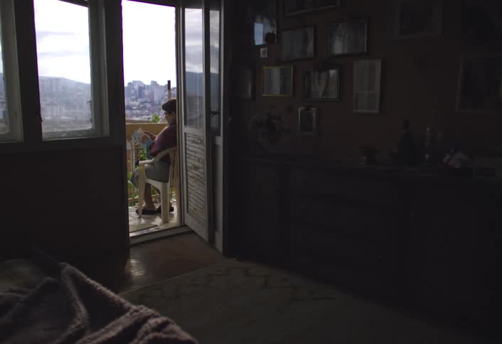 A person is sitting on a balcony chair reading with a cityscape and mountains in the background while the interior of the room is dimly lit