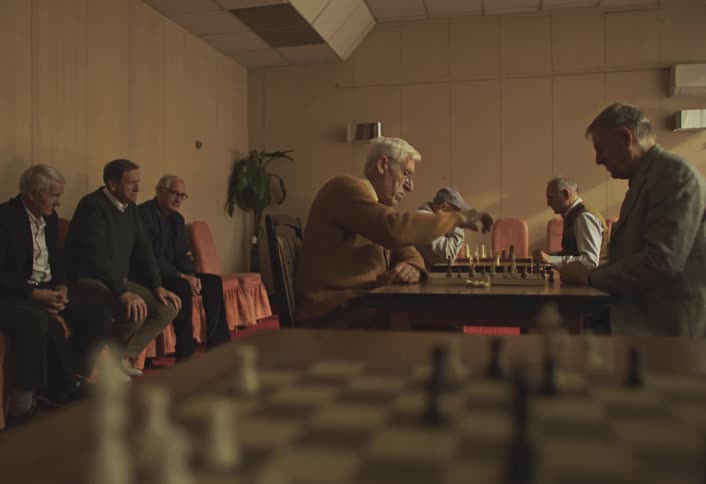 Elderly men play chess in a dimly lit room while others sit in the background watching