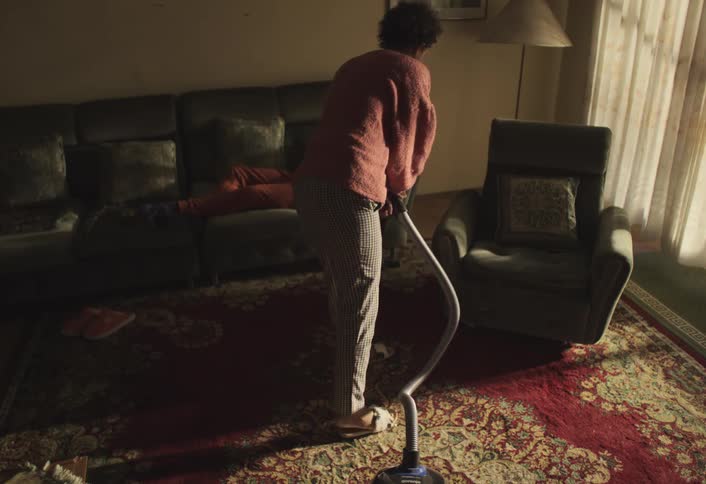 Woman vacuums the floor while another woman lying on the sofa throws an object into the air in a room lit by natural light
