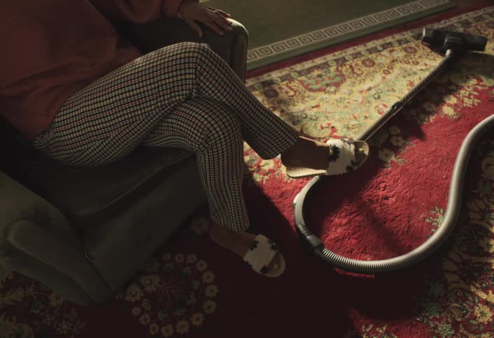 Woman sits in an armchair with her legs crossed next to a vacuum cleaner on a carpet