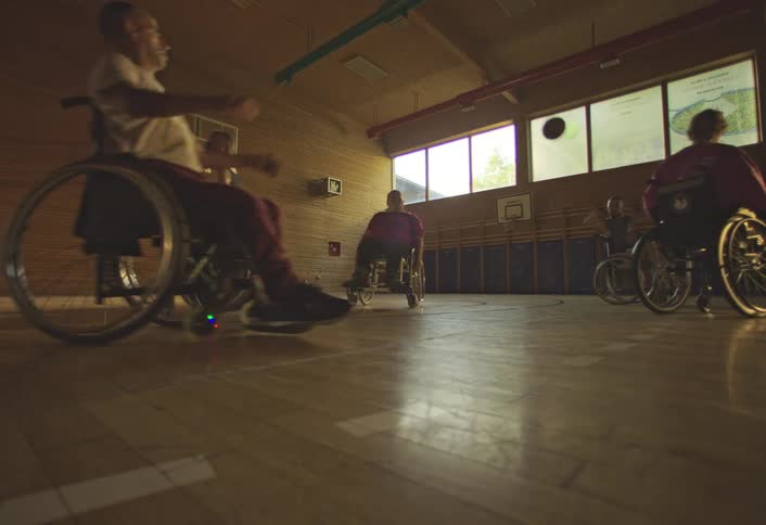 People in wheelchairs play basketball in a gymnasium lit by natural light from windows