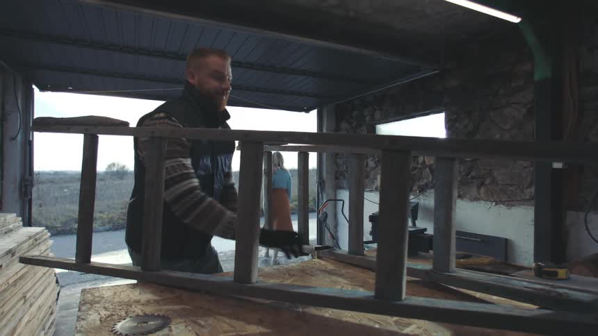 A person is working in a workshop holding a wooden ladder on a workbench with various tools and materials visible around
