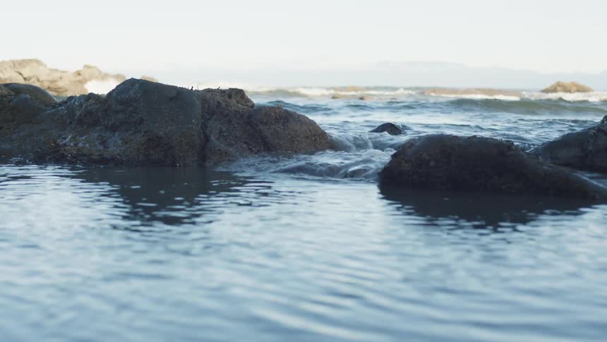 Water flows through a gap between coastal rocks as distant waves break against the shore under a clear sky