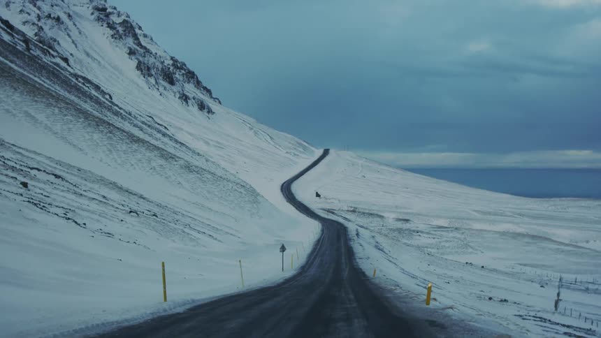 A winding mountain road stretches up a snowy slope flanked by bright yellow marker posts on both sides