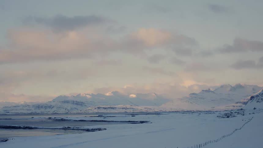 A vast snow covered landscape stretches towards a misty mountain range in the distance with a fence line receding into the foreground