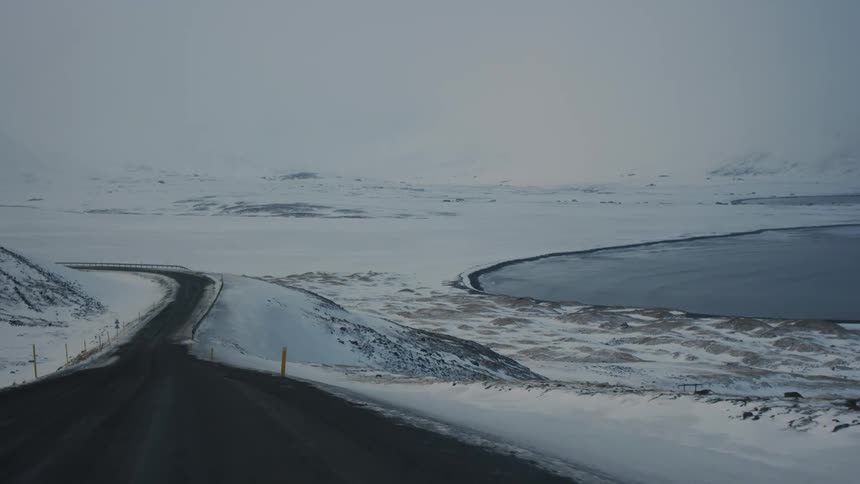A mountain road winds down a snowy slope revealing a tranquil lake below and bright yellow markers along the way