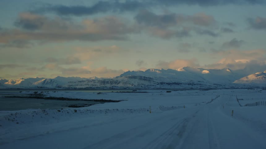 A vast snow covered landscape extends towards a distant mountain range under a cloudy sky with a road and fence line leading the eye into the scene suggesting a remote and wintry environment