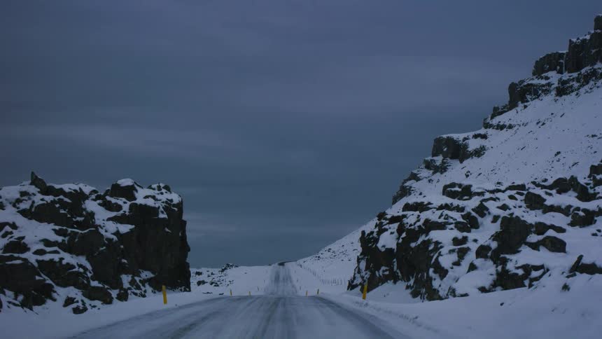 A mountain road passes through a rocky opening in a snowy slope before descending downhill under a moody winter sky