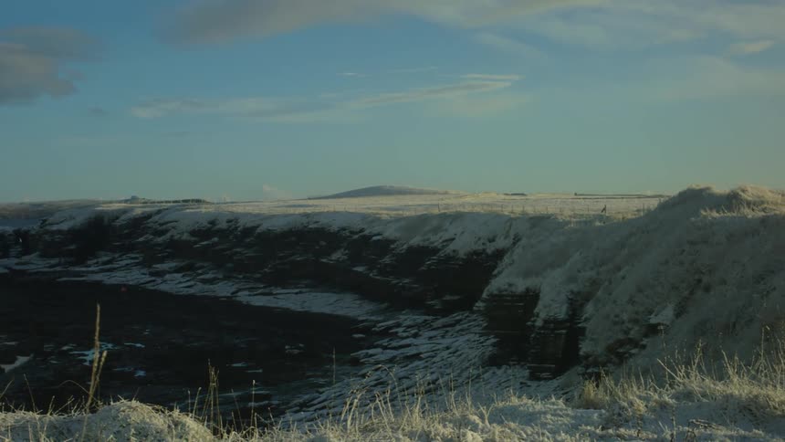 A vast snow covered landscape stretches under a pale sky featuring a dark rocky cliff receding into the distance