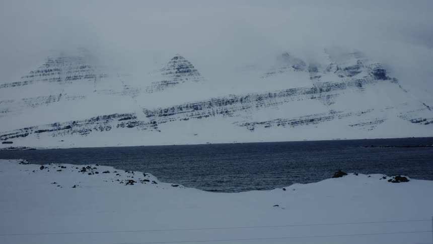 Snow covered ground beside a lake at the bottom of a snow covered mountain