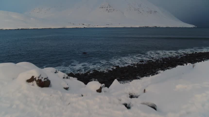 A rugged snow covered coastline near the churning lake under an overcast sky with waves crashing against the rocky shore and a vast white mountain looming in the background