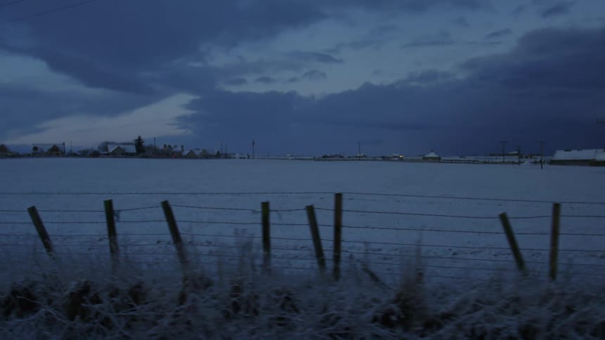 A snowcovered field stretches towards a line of trees under a dark dramatic sky