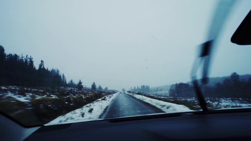 A snowy treelined road disappears into the foggy distance as seen through a car windshield with active wipers