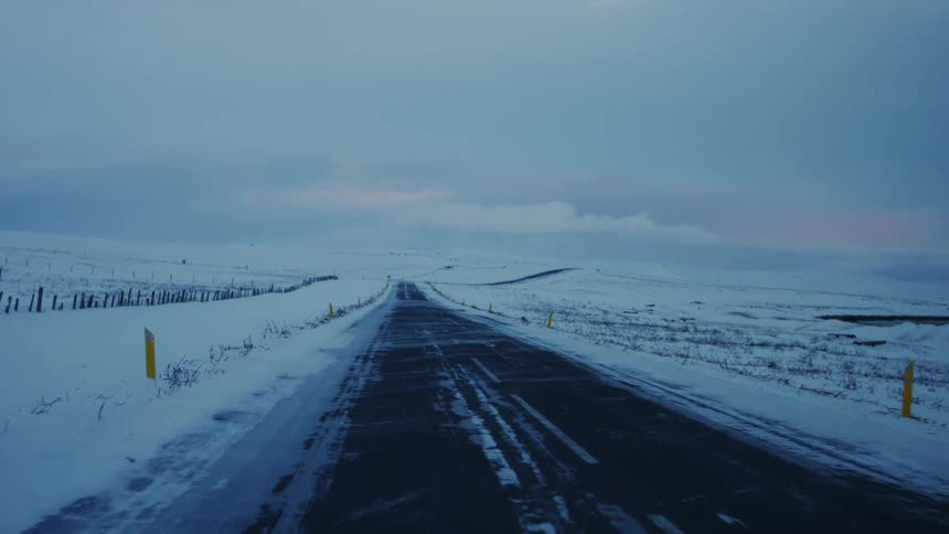 The view from within a vehicle travels down a frozen road lined with yellow markers passing through snowy farmland under an overcast sky