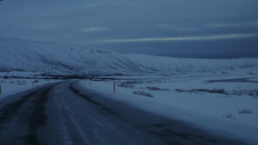An icy mountain road stretches through a snowy landscape with yellow markers and a distant river visible under an overcast sky