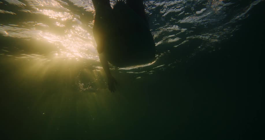 A silhouette of a person floating on a surfboard is illuminated by sunlight filtering through the surface