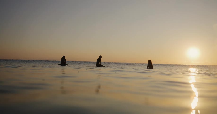 Three people are sitting on surfboards in the sea during sunset with reflections of the sun on the water