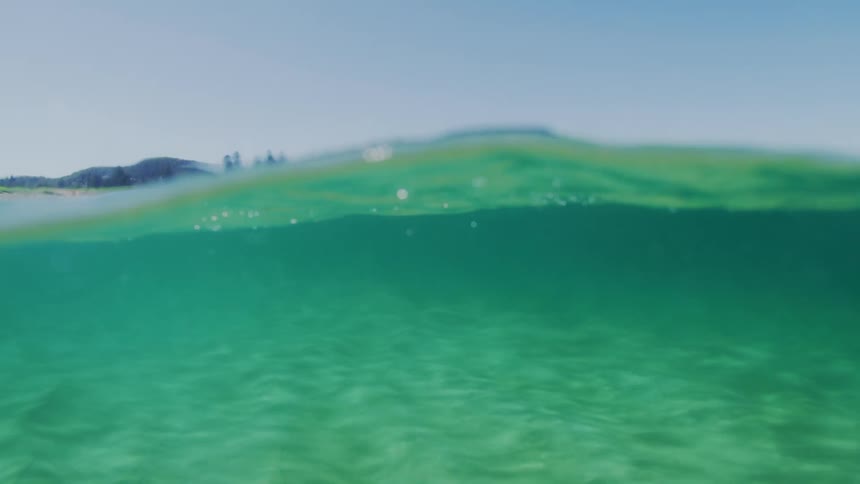 A split view of a clear greenishblue ocean with a sandy bottom and a distant coastline featuring a rocky cliff and trees under a clear sky