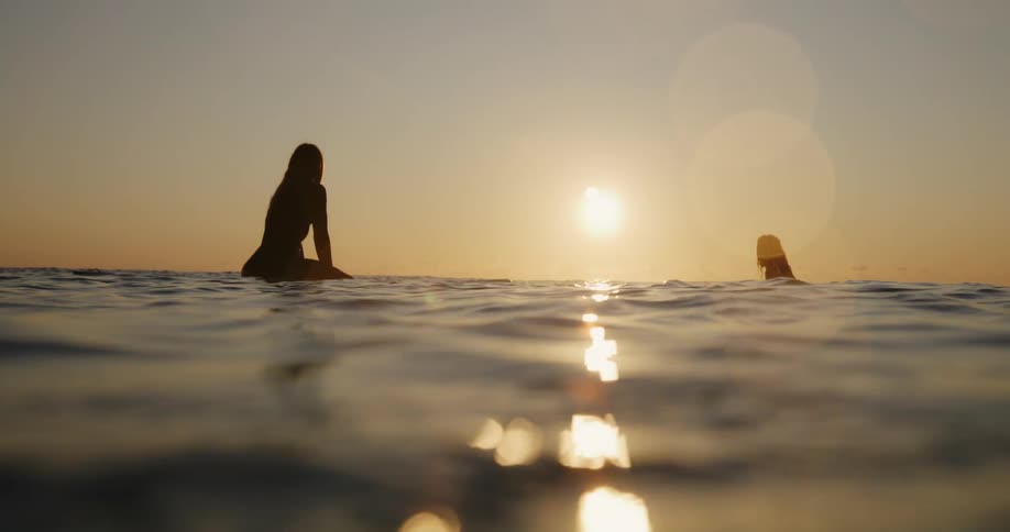 Two female surfers sit silhouetted on their surfboards in the ocean at sunset