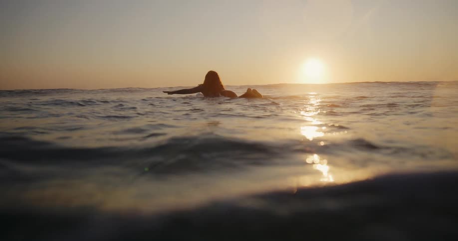 Silhouette of a woman lying on a surfboard floats in the sea as she watches the sun set on the horizon