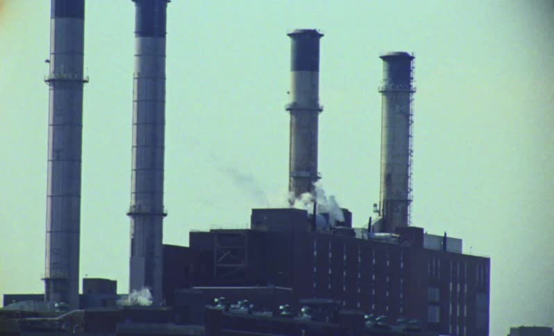 Four tall smokestacks rise from a dark industrial building emitting white plumes of smoke against a light blue sky
