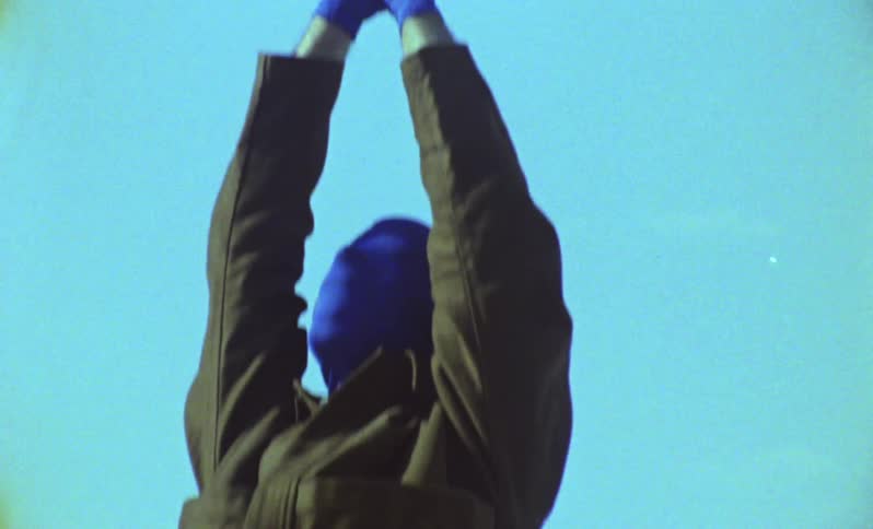Person wearing gloves and a mask covering their face with their hands clasped together and standing on a rock at the edge of a lake