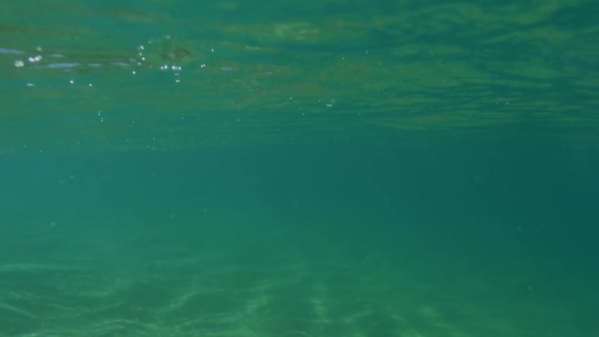The point of view from just above the surface of a clear turquoise sea with the blurred horizon line dividing the frame and a distant rocky coastline barely visible under a bright sky