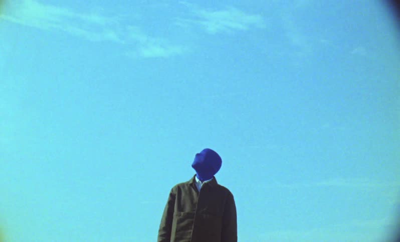 Person wearing a dark coat and blue hood completely covering the face standing against a clear sky with few clouds