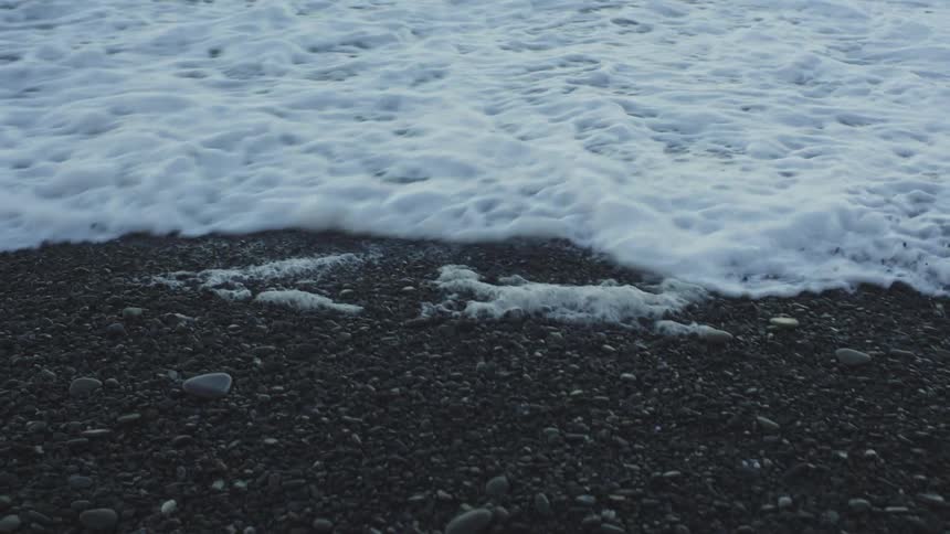 Waves foam as they flow onto a beach covered by dark pebbles