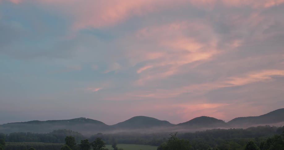 A tranquil misty landscape unfolds with rolling hills and valleys shrouded in a soft fog punctuated by lush green forests and a hint of sunlight breaking through the clouds