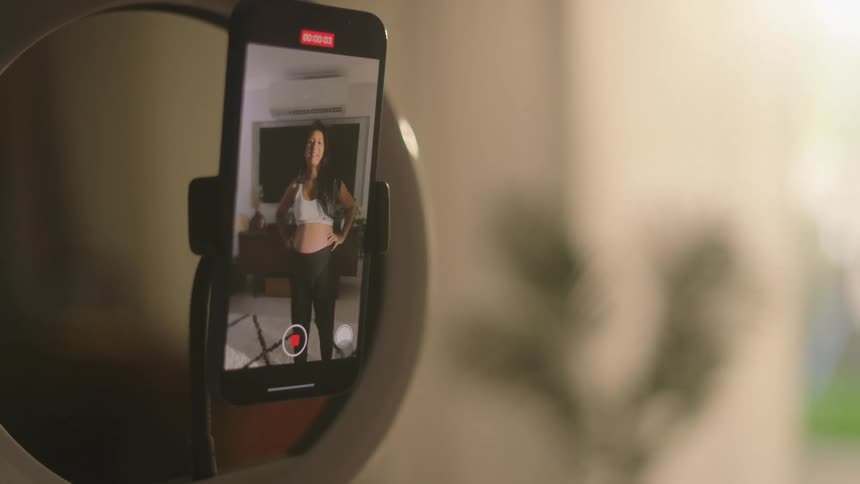 A pregnant woman in a white tank top is recording or streaming from a cozy living room