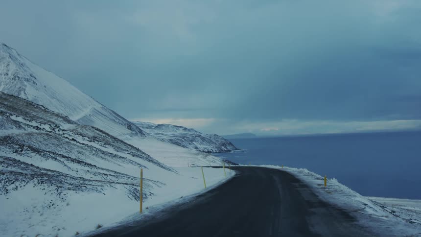A winding road cuts through a snow covered mountain landscape leading towards a calm ocean bay under an overcast sky