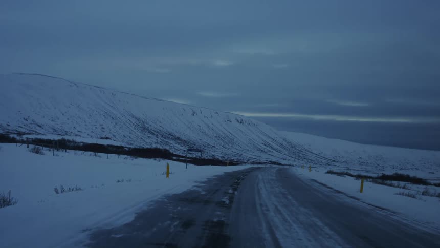 A wet snow covered road winds through a desolate wintry landscape leading towards a dark imposing mountain range under a heavy overcast sky