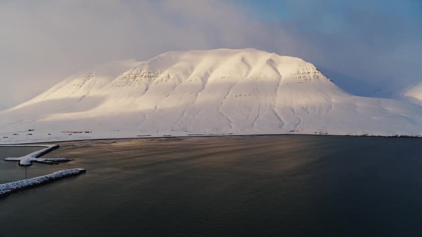 A snow covered mountain slopes down to a dark harbor contrasting the stark white of the snow with the deep blue water and the faint hazy sky above
