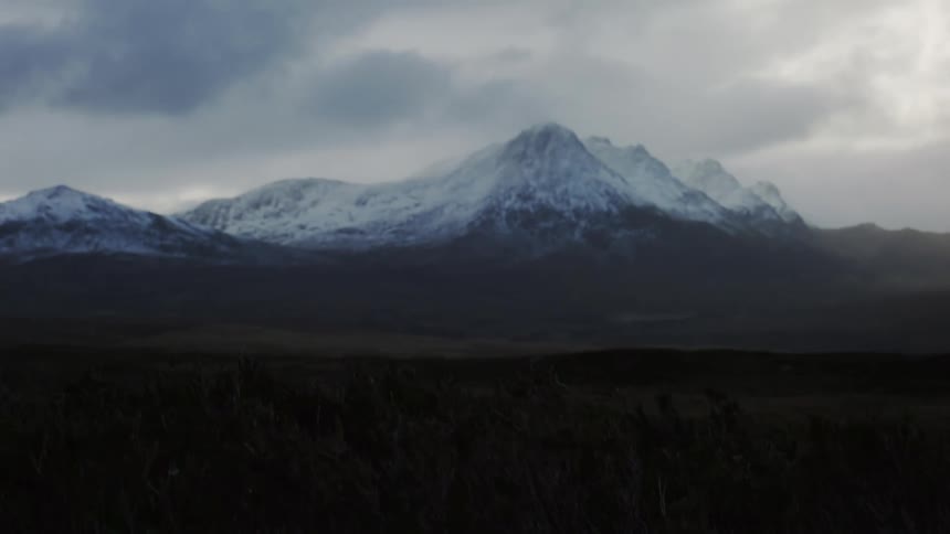 Snow capped mountains rise against a moody overcast sky their dark forms contrasting with the patches of white snow and the somber foreground