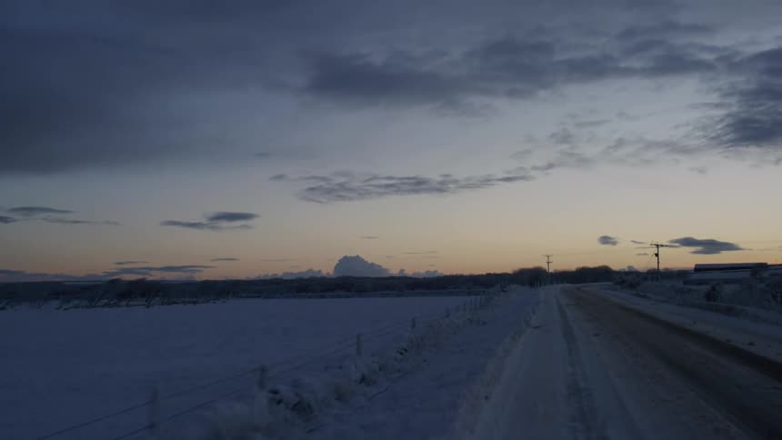 A snow covered landscape stretches into the distance under a twilight sky featuring a road leading towards a silhouetted horizon with power lines