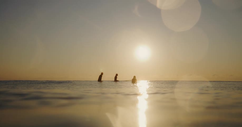 Three figures stand silhouetted in the ocean at sunset their surfboards catching the light as they face the horizon