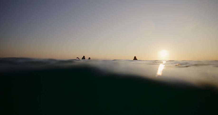 Silhouettes of surfers floating in the sea at dusk with the surface of the water reflecting the golden light of the sun near the horizon