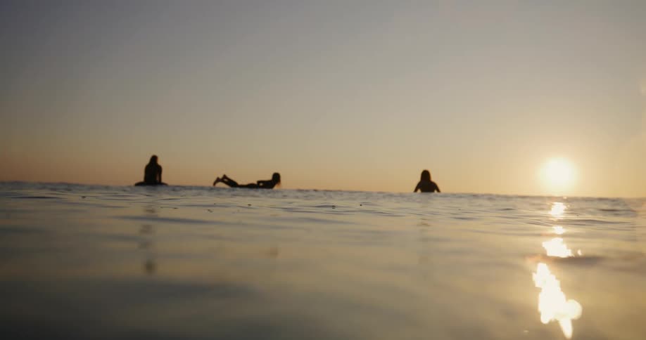 Silhouette of three surfers in a calm sea with the sun on the horizon