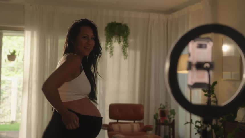 A pregnant woman is filming herself with a ring light and smartphone in a living room setting