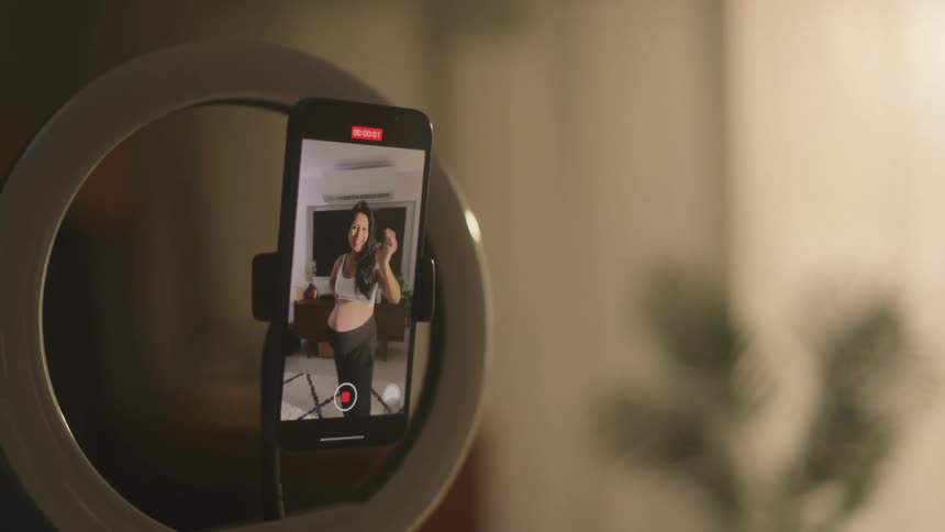A smartphone mounted in a ring light holder displays a womans face as she touches the screen to begin recording a video of her pregnant belly