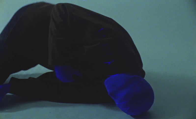 A model with a blue head and blue hands dressed in a dark suit lies on the ground while moving during a photo session in a studio