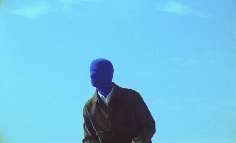 Man wears a blue face and head covering while posing outdoors with the city skyline in the background
