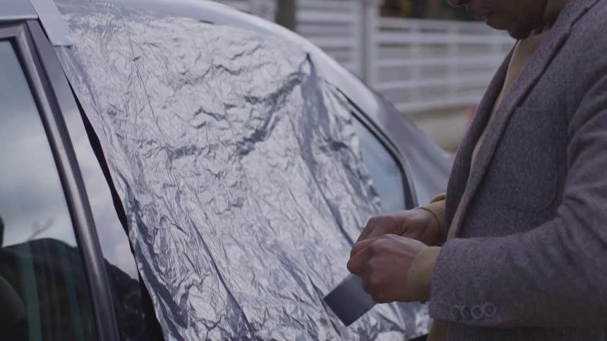 A man tapes a piece of plastic to a broken car window