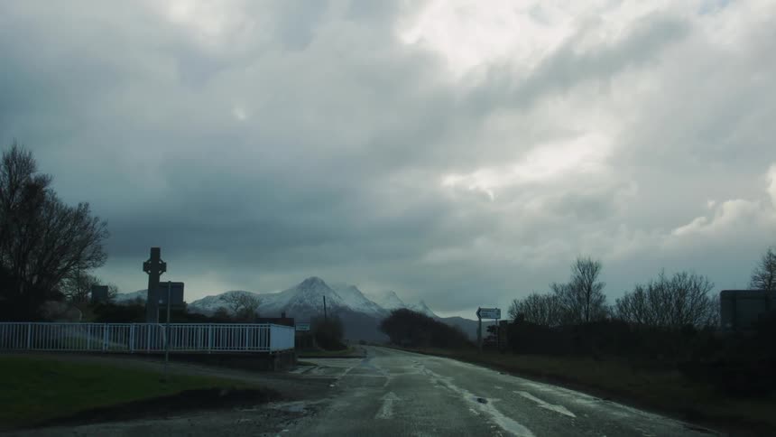 A wet road stretches toward distant snow capped mountains under a cloudy sky framed by a roadside graveyard and trees