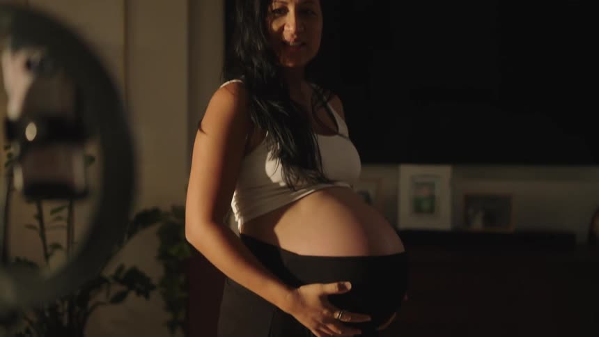 A pregnant woman in a white tank top is recording or streaming from a cozy living room