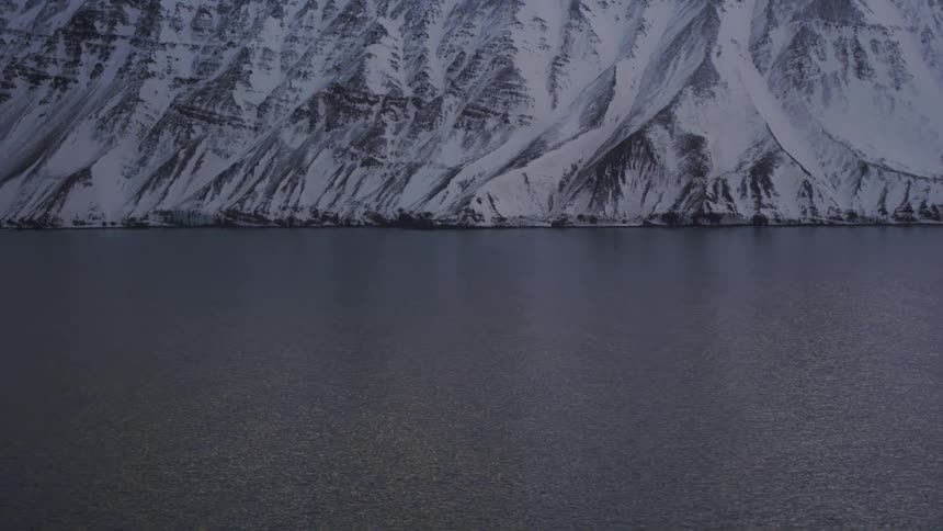 A snowcovered mountain slope rises sharply from a dark still body of water under a cloudy sky viewed from a moving vehicle