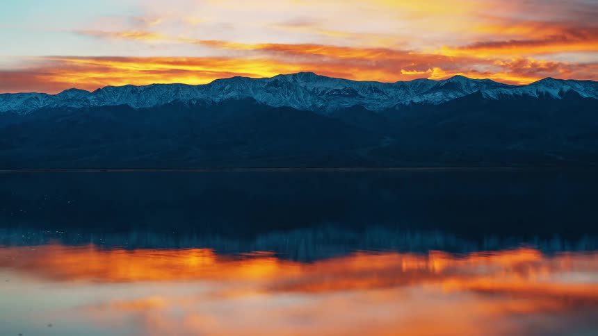 A vibrant sunset sky ablaze with orange and yellow hues is reflected in the still water of a lake mirroring the dark silhouette of distant mountains